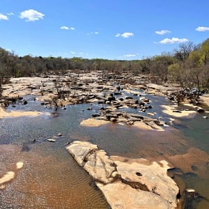 Shallow river over broad, flat rocks, bordered by woods beneath a bright blue sky.