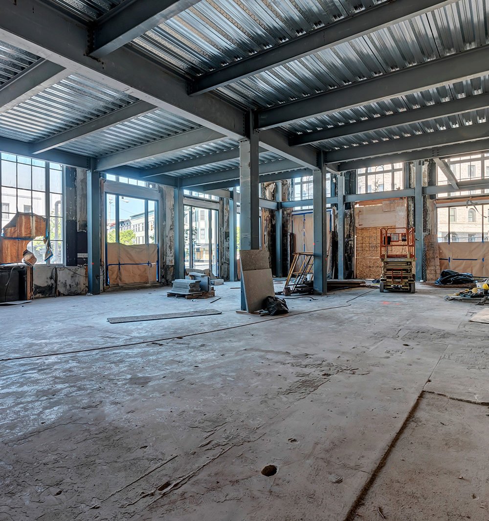 Unfinished interior with exposed steel framing, a concrete floor, and construction equipment by large windows.