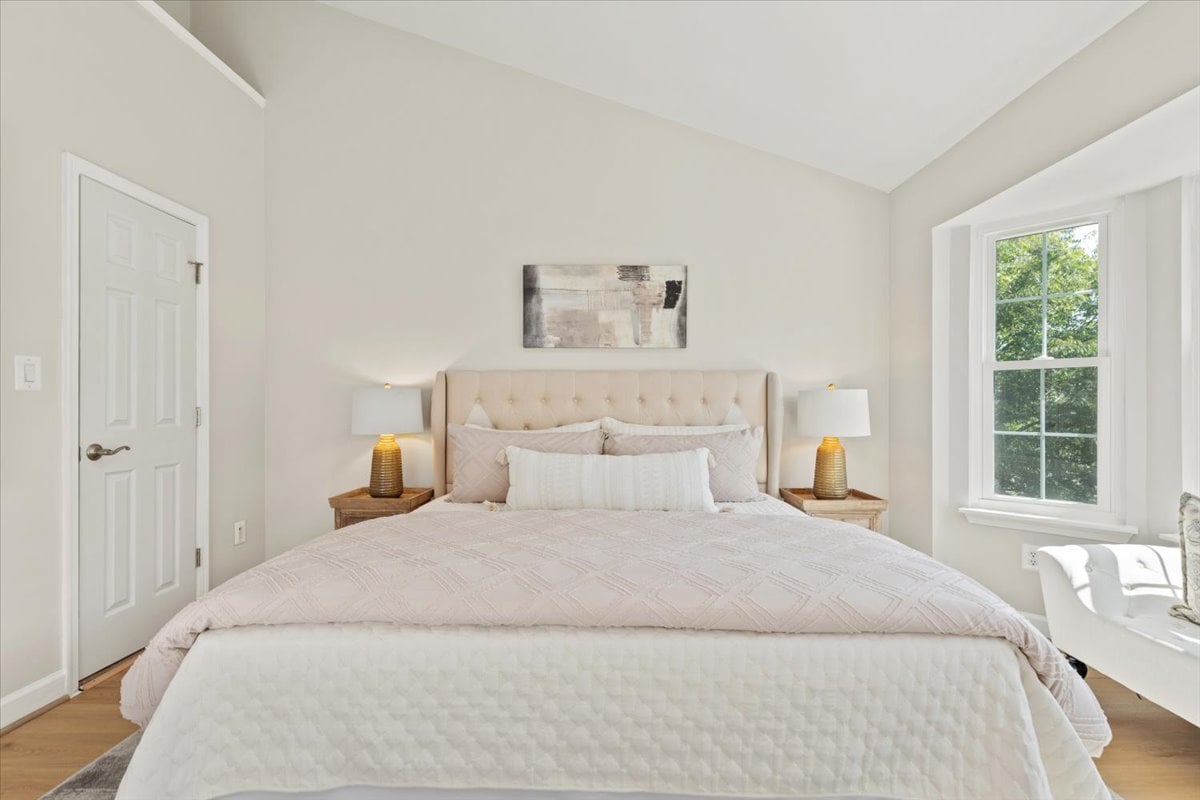 Staged bedroom with a large bed, tufted headboard, matching lamps, and a window overlooking greenery.