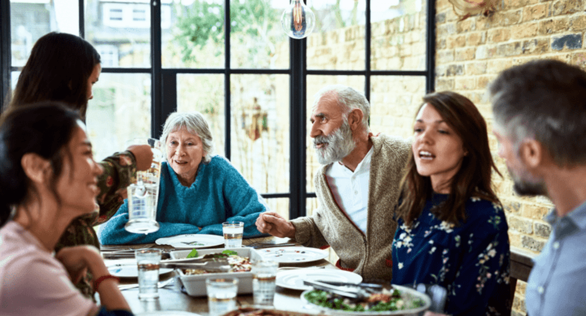 Adults of different generations share a meal around a dining table in a bright, brick-walled room.