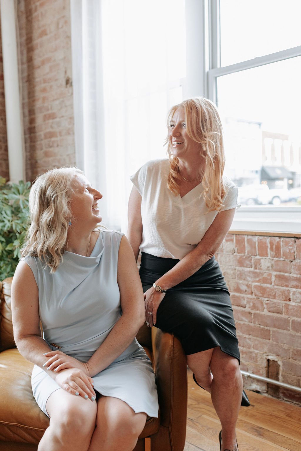 Two women smiling together beside a large window in a brick-walled interior.