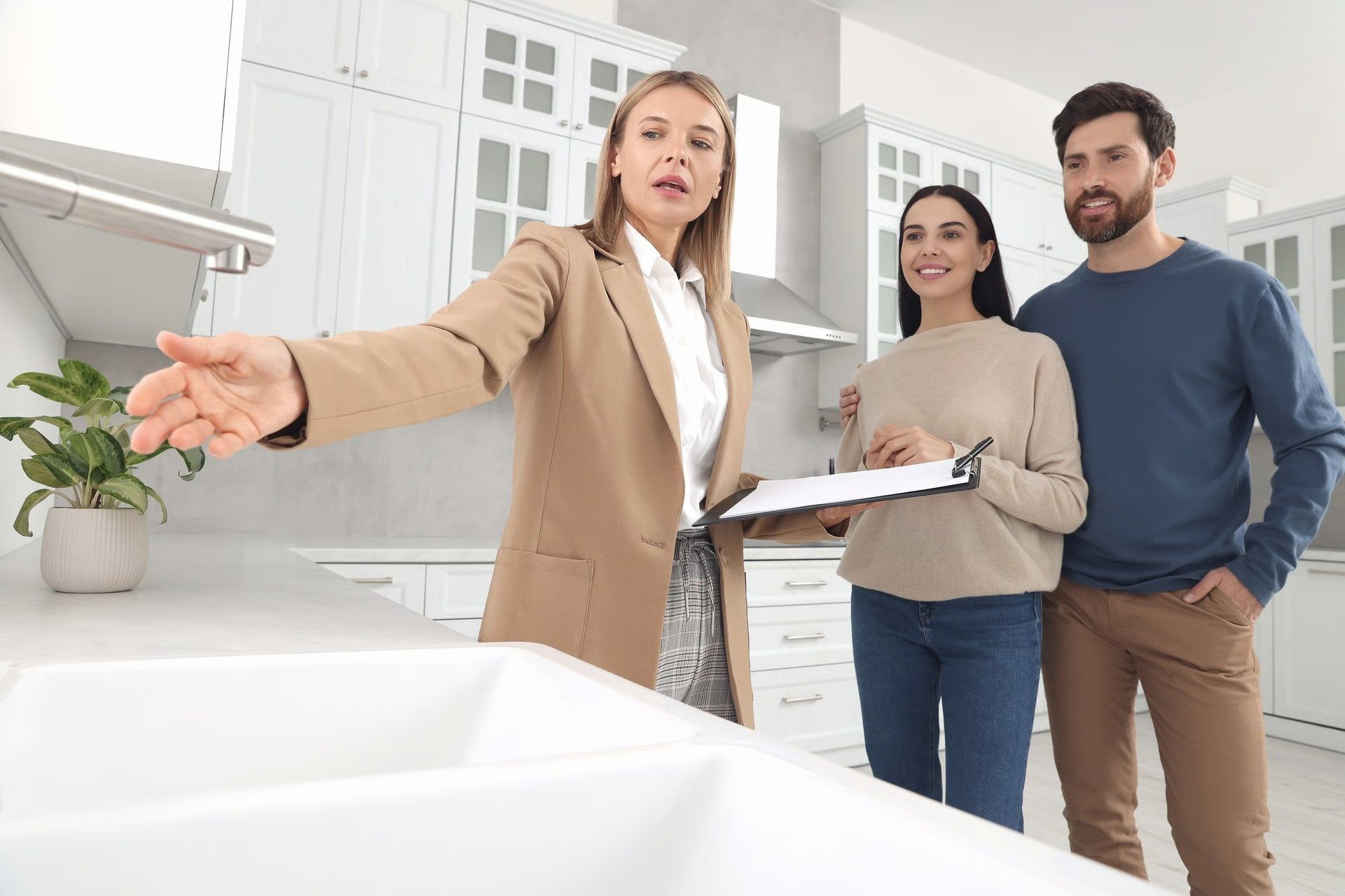 Real estate agent showing a white kitchen to a couple, with the woman holding a clipboard.