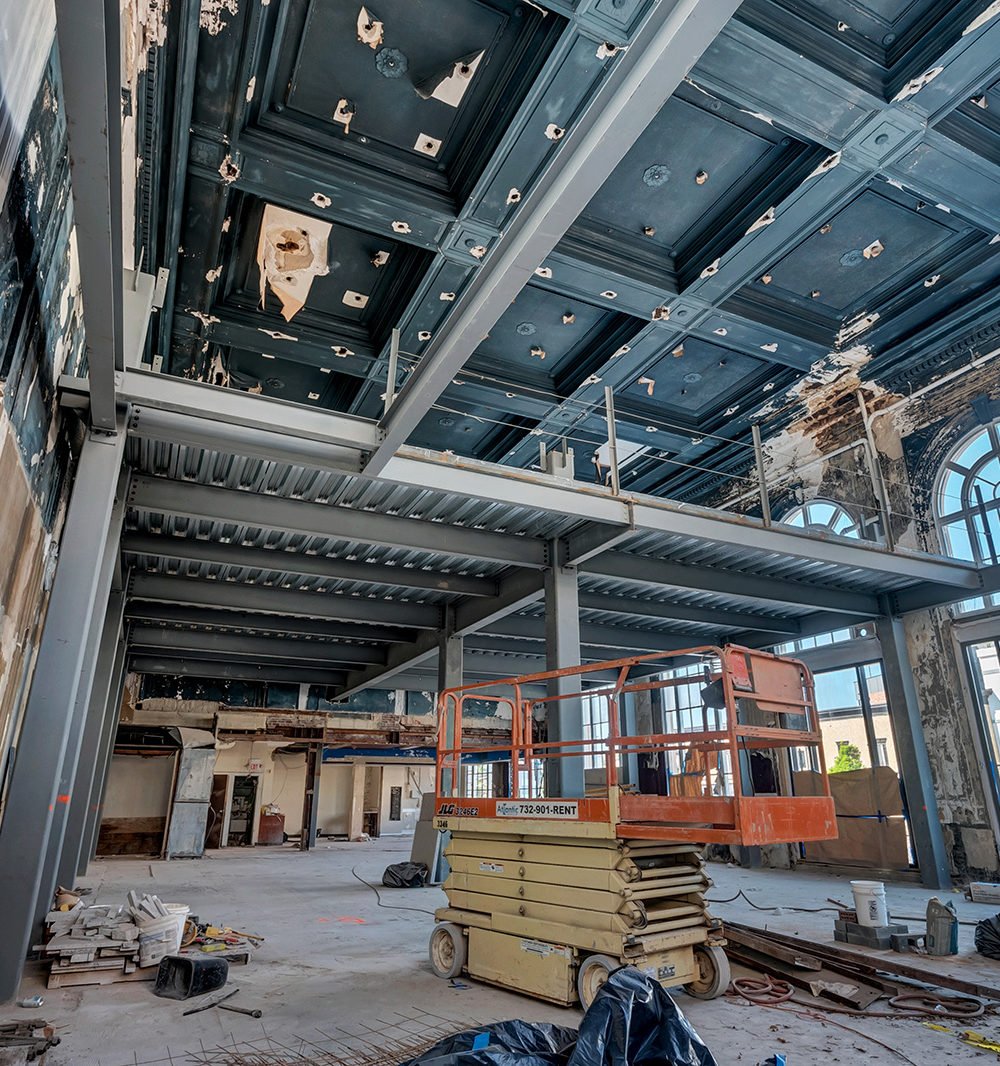 Historic interior under renovation with a blue coffered ceiling, steel mezzanine, and orange scissor lift.
