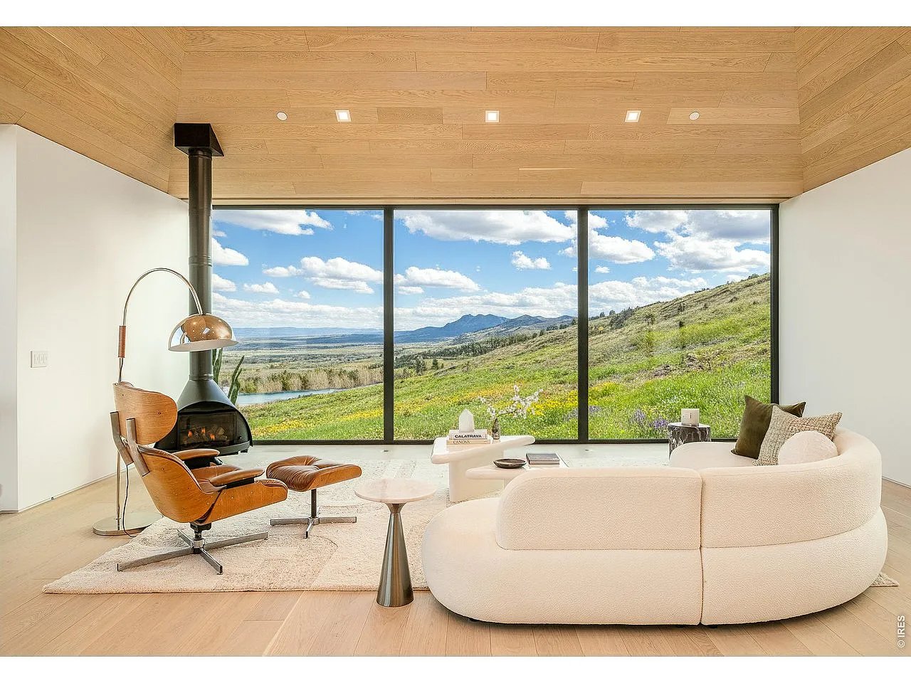 Modern living room with a curved white sofa and wood stove overlooking a lake, green hillside, and distant mountains.