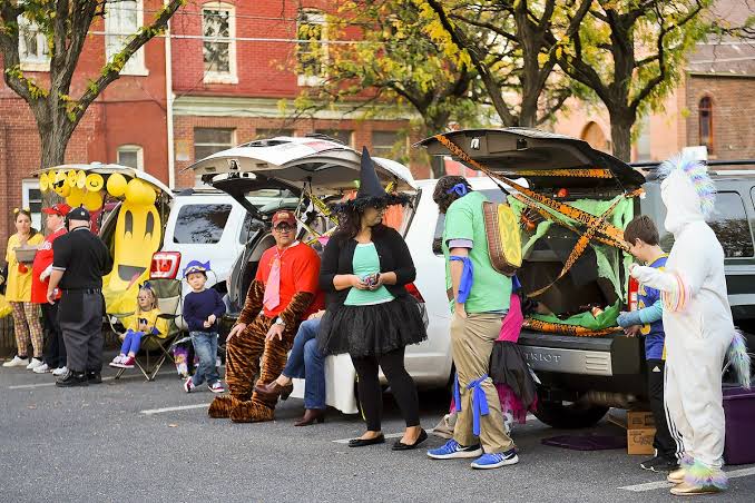 People in Halloween costumes gather beside decorated cars with open trunks on a tree-lined street.