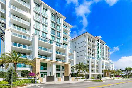 Modern white condominium buildings with glass balconies beside a palm-lined street.