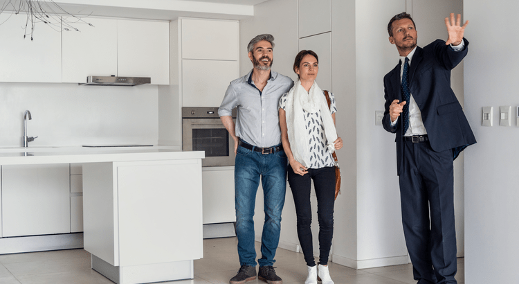 A couple stands in a modern kitchen, attentively listening to a real estate agent in a suit pointing ahead. The space is bright, conveying optimism.
