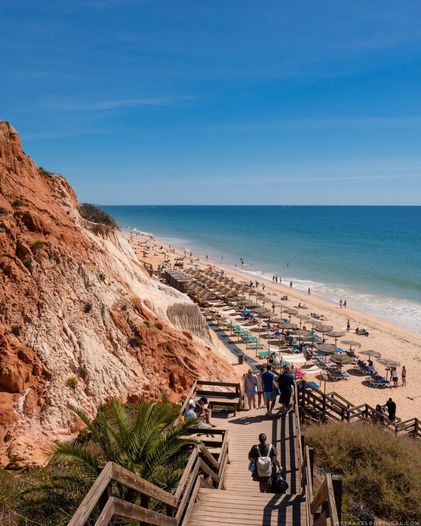 A wooden staircase leading down to Praia da Falesia, where parasols line the shore below eroded red cliffs