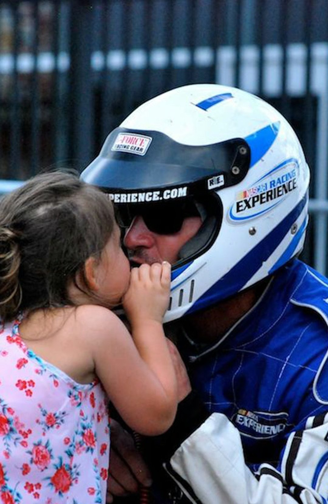 A young girl kisses a race car driver in a white-and-blue helmet and racing suit.