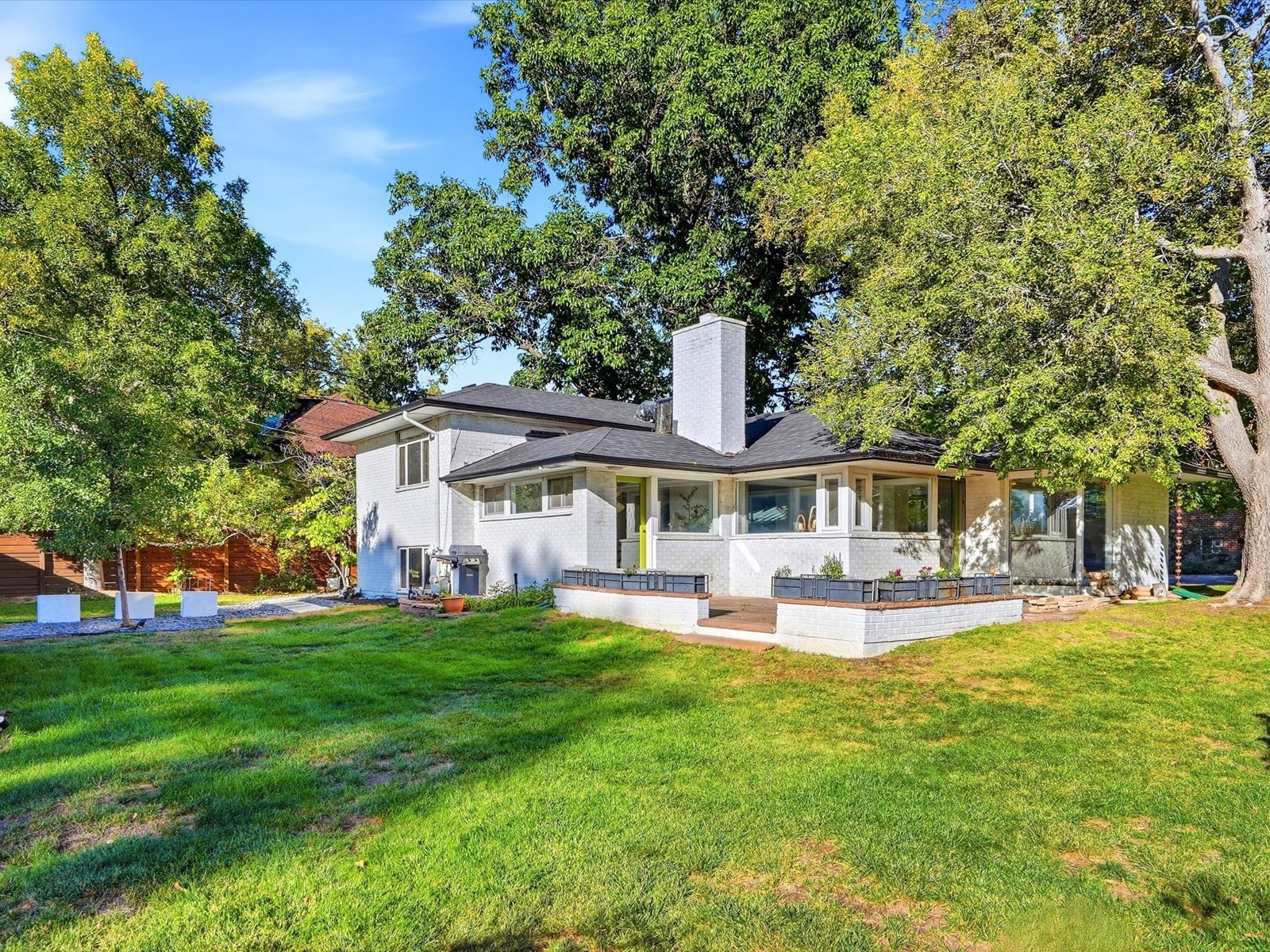 White-brick mid-century modern home with a tall chimney, raised planters, and a tree-shaded lawn.
