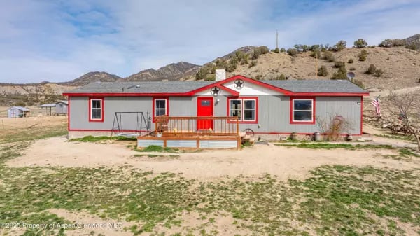 Gray single-story home with red trim and a small front deck, backed by dry hills.