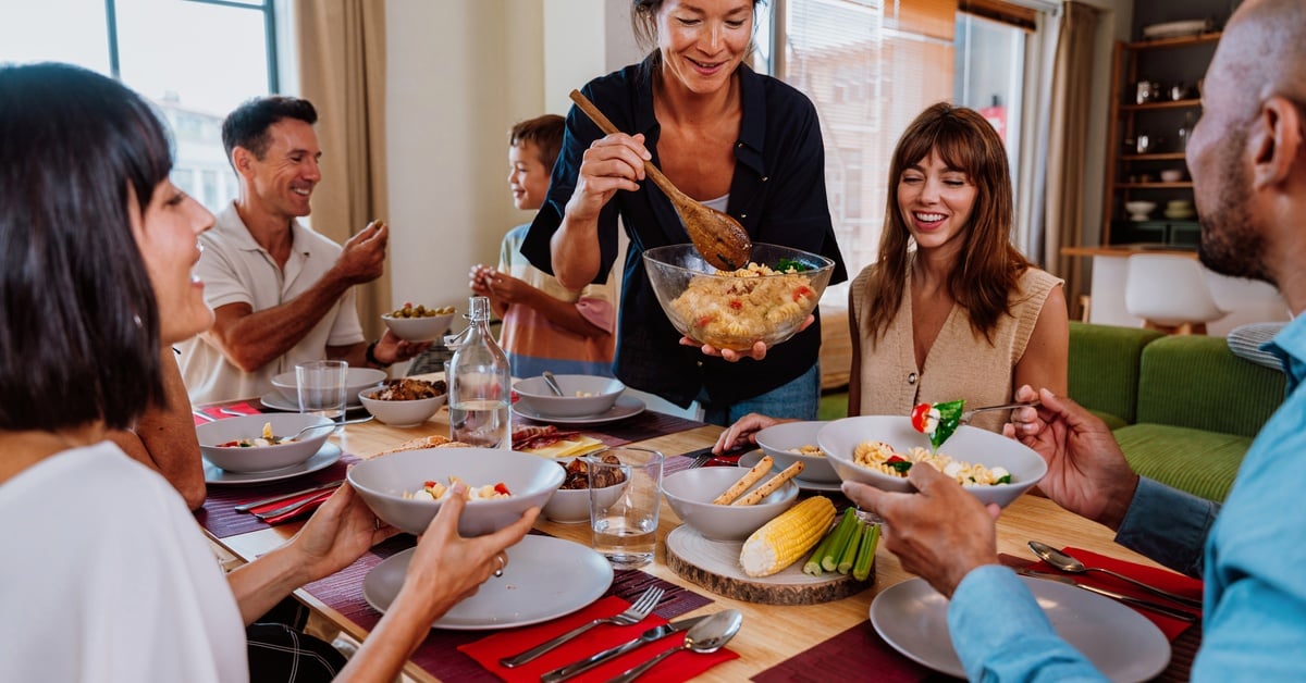 A multigenerational family sitting at a large table eating dinner together, with an older woman ladling out food on dishes.