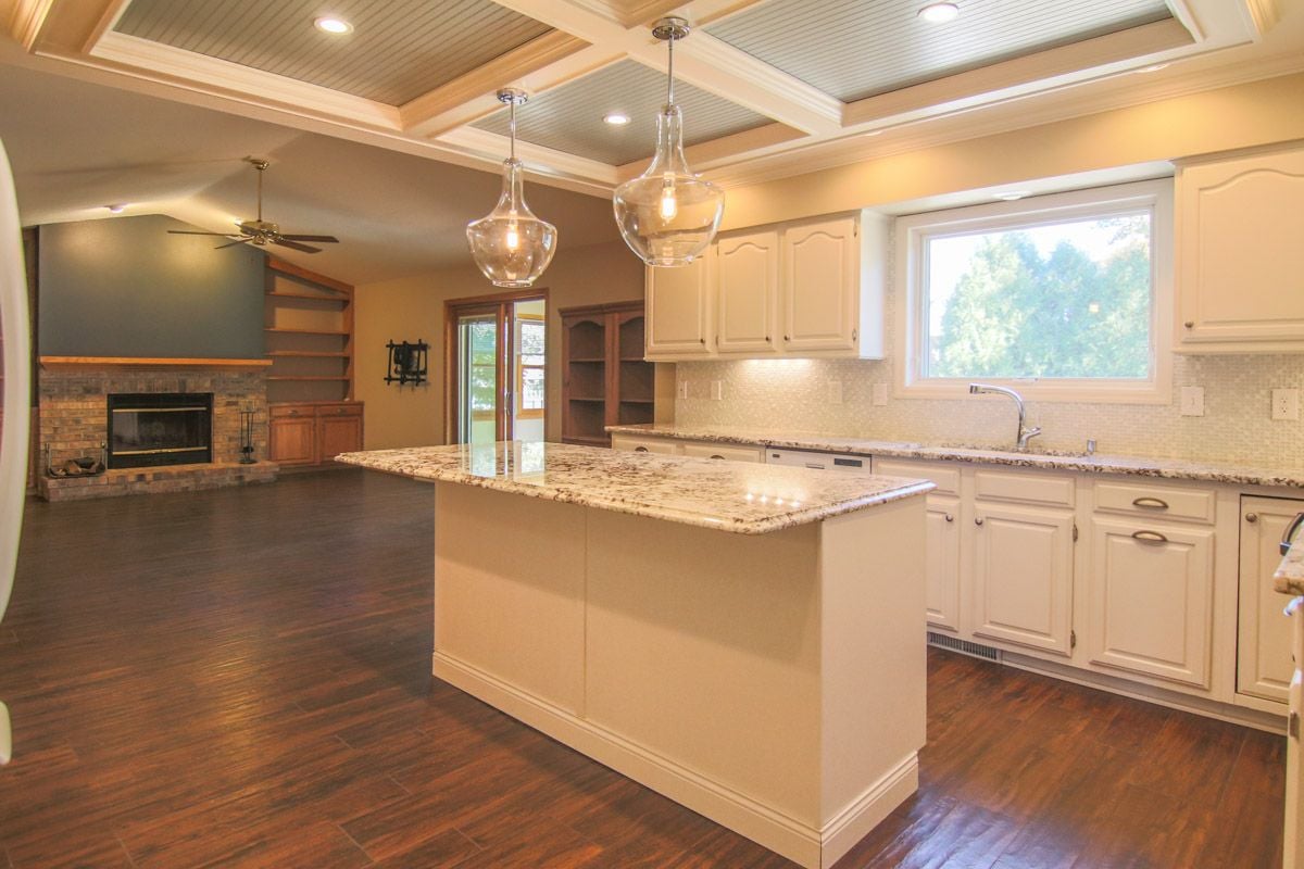 Open-plan kitchen with a granite island, white cabinets, coffered ceiling, and adjoining stone fireplace.