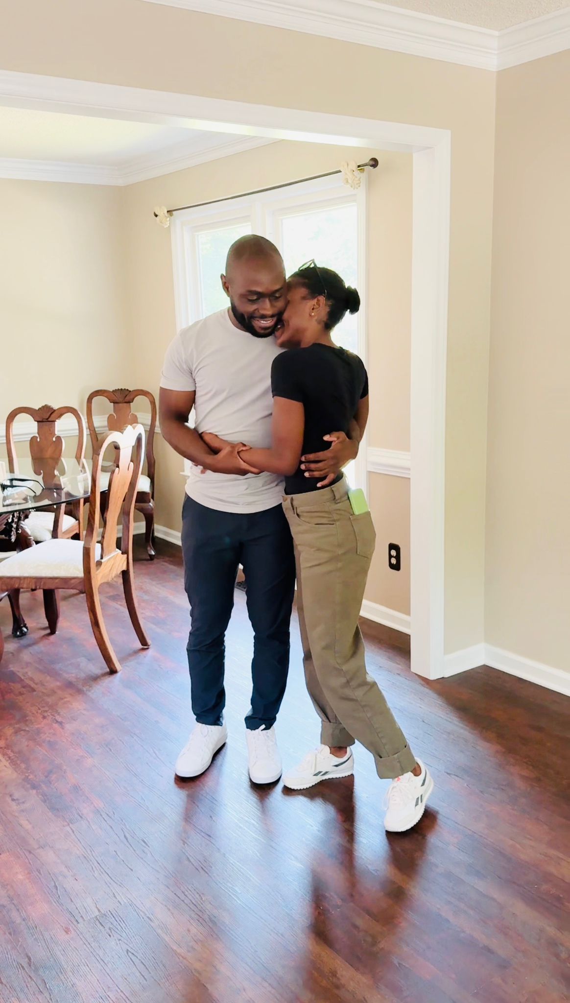 A happy young couple embraces and smiles inside their new home with hardwood floors and a bright dining area in the background.