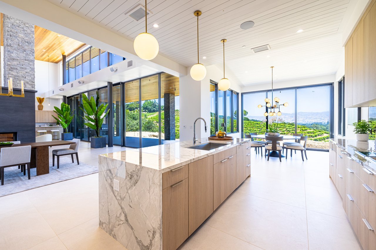 Modern kitchen with a marble island and expansive windows overlooking vineyard rows and rolling hills.