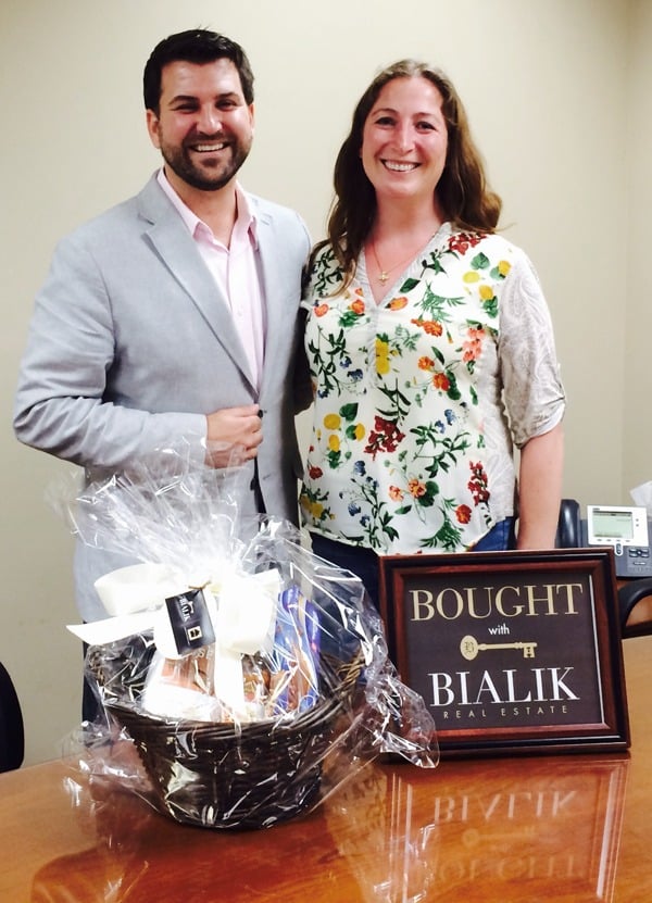 A smiling couple pose with a gift basket and a framed Bialik Real Estate closing sign.