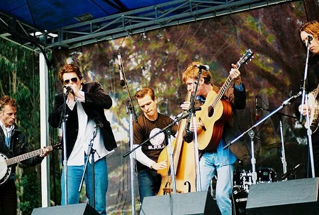 Band performing on outdoor stage with four musicians playing acoustic guitar, upright bass, and banjo while lead singer with sunglasses holds microphone, backed by colorful projected backdrop