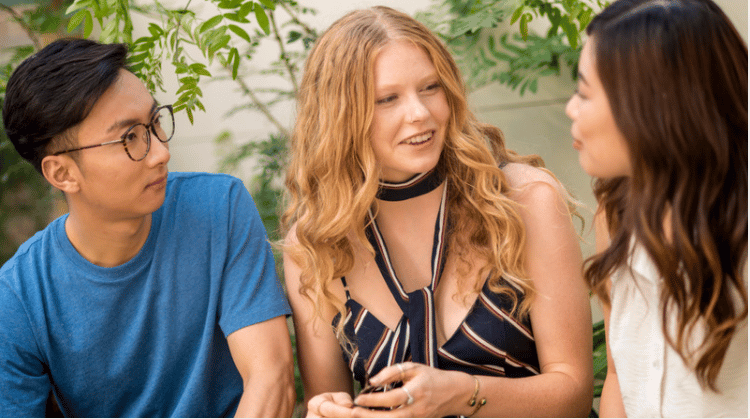 Three teenagers talking together outdoors beneath leafy branches.