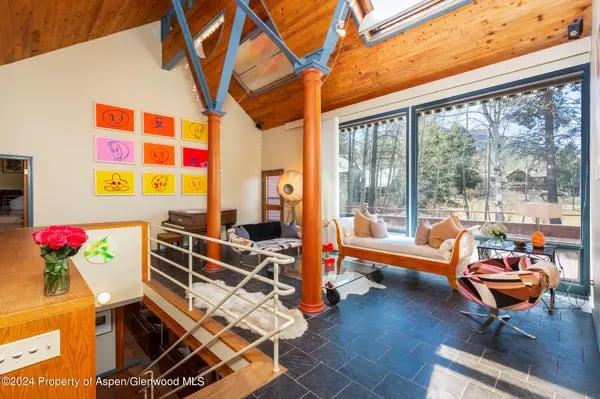 Loft living room with a vaulted wood ceiling, orange support column, and large windows overlooking trees.