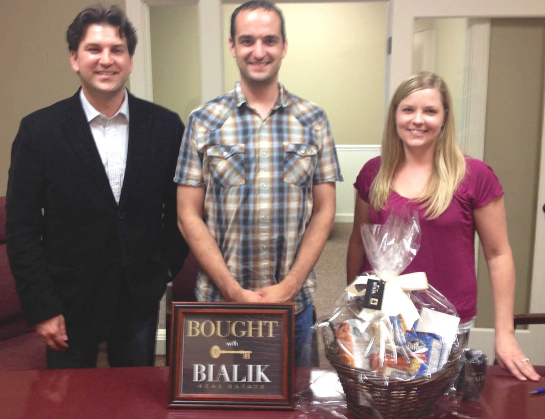 Two men and a woman pose with a Bialik Real Estate “BOUGHT” sign and gift basket.