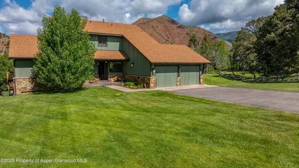 Green two-story home with an attached garage, broad lawn, and mountains behind it.