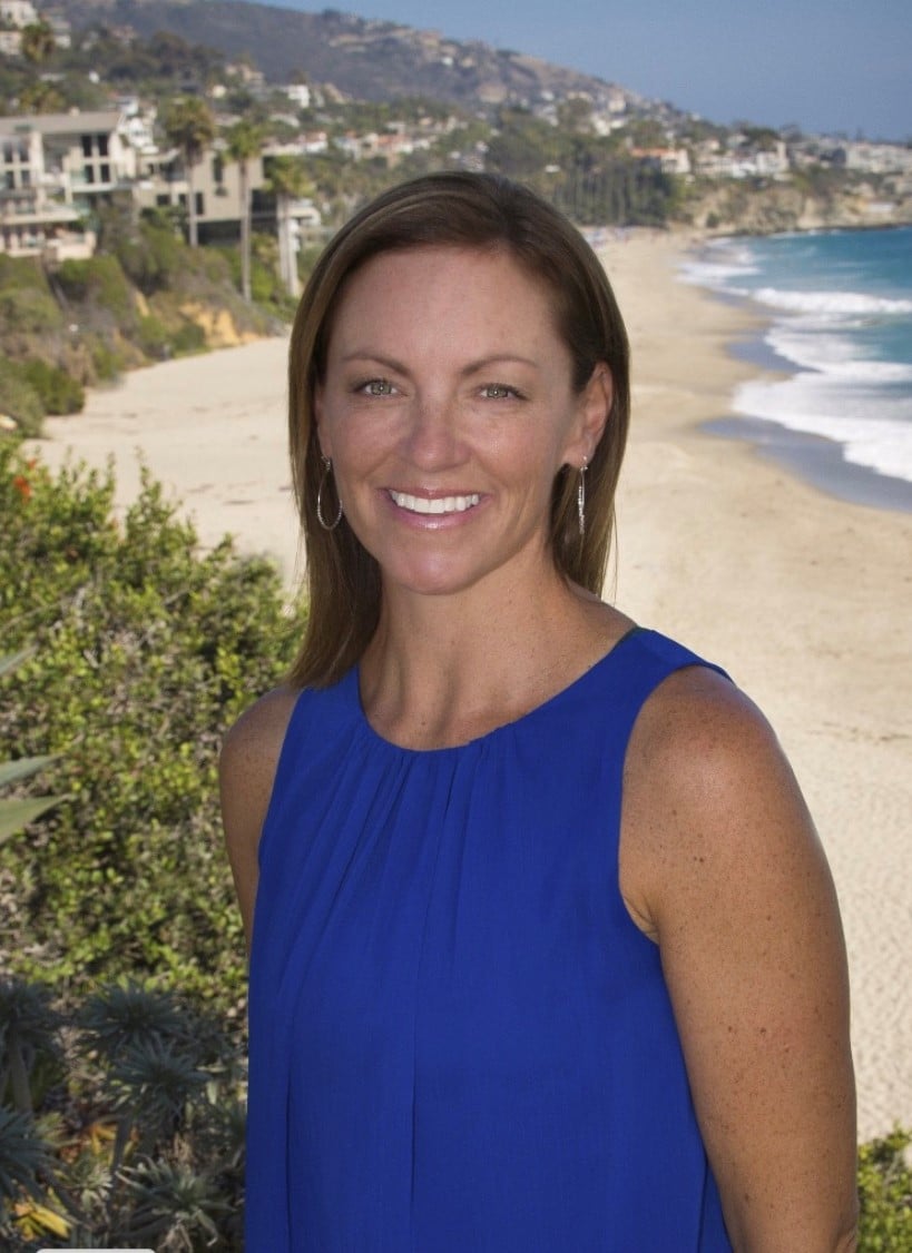 Smiling woman in a blue sleeveless top, with a beach and hillside homes behind her.