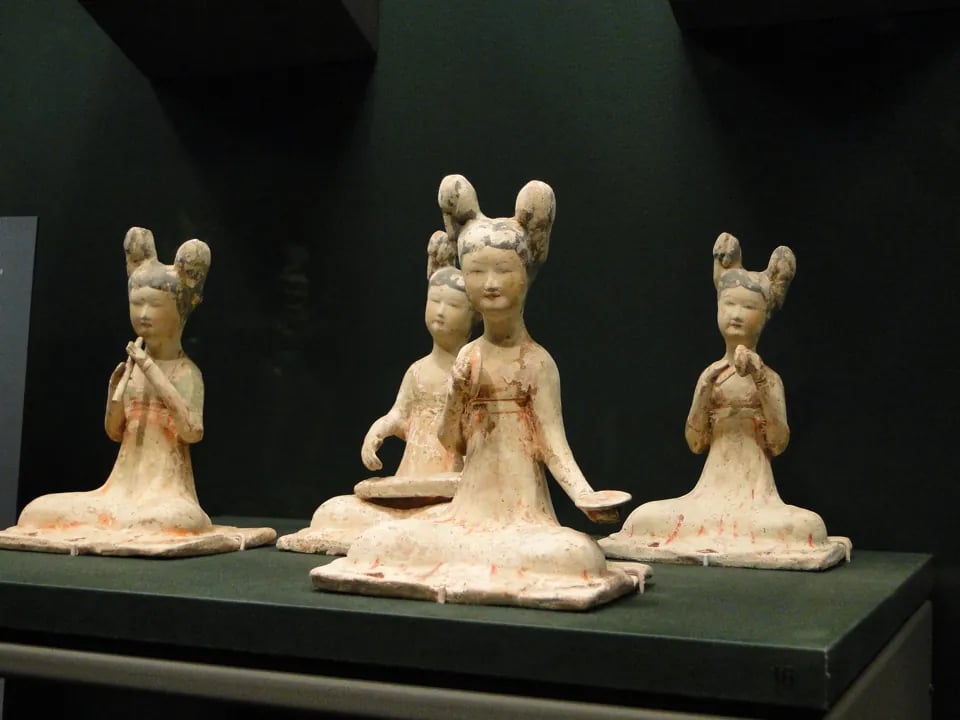 Four ancient Chinese ceramic figurines of seated female musicians with elaborate double-bun hairstyles displayed at the Asian Art Museum.