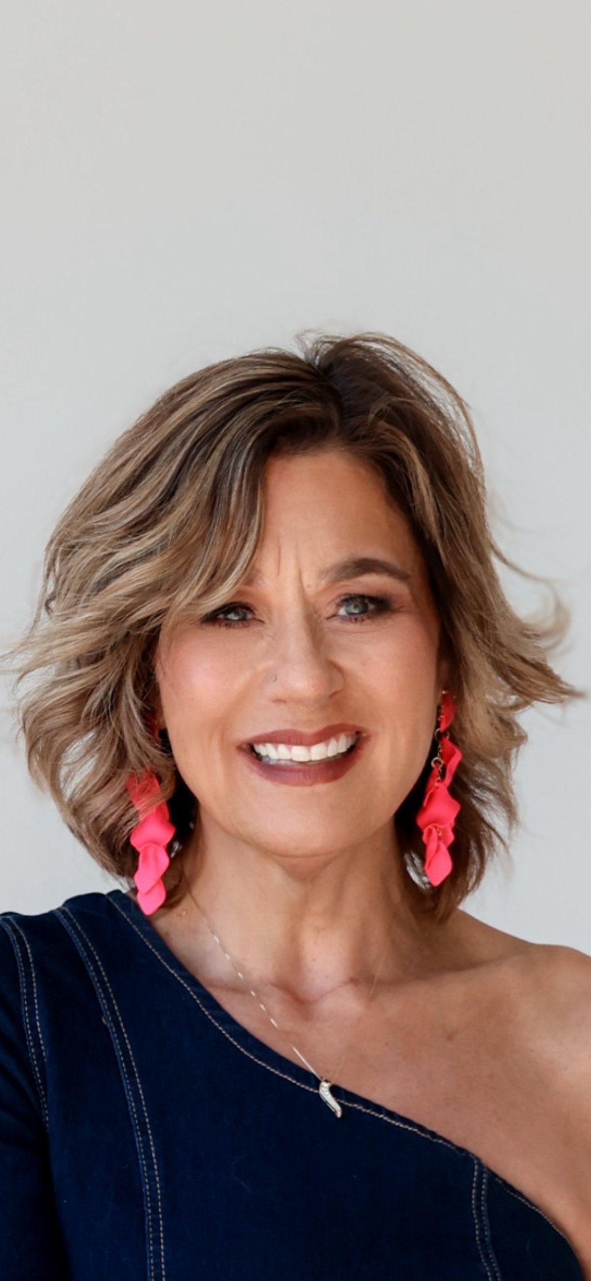 Kim Craun smiling in a navy one-shoulder top and bright pink earrings.