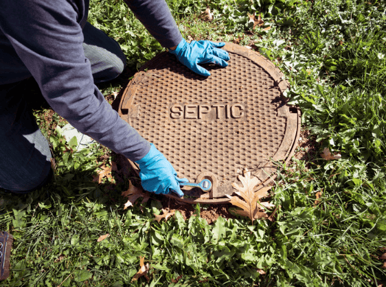 A worker checks a round septic tank cover in a grassy yard.
