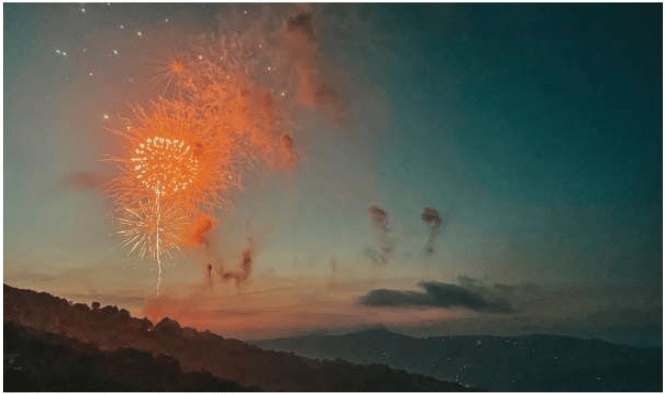 Fireworks burst above layered mountain ridges at dusk.