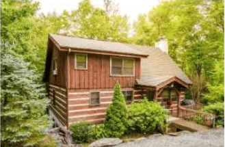 Rustic log cabin with a covered porch and stone chimney surrounded by lush trees.