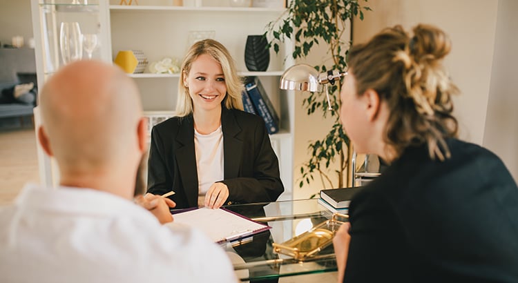 A woman reviews paperwork with two clients across a glass desk in a bright office.