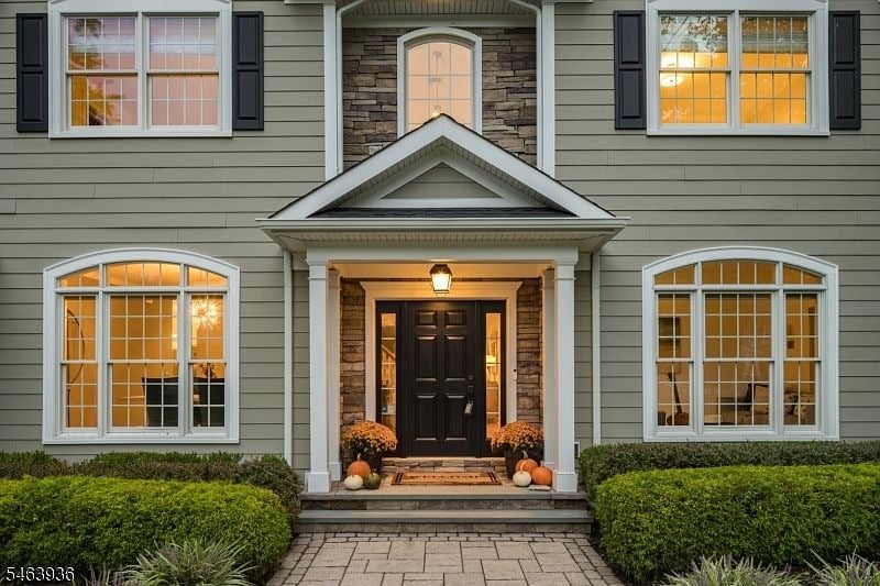 Two-story gray house with stone entry, glowing windows, and pumpkins by the front door.