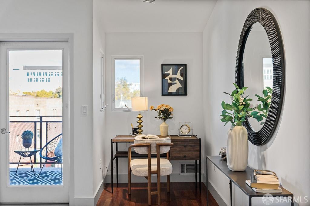 Staged home office nook with a dark wood desk, upholstered chair, and oval mirror beside a balcony door.