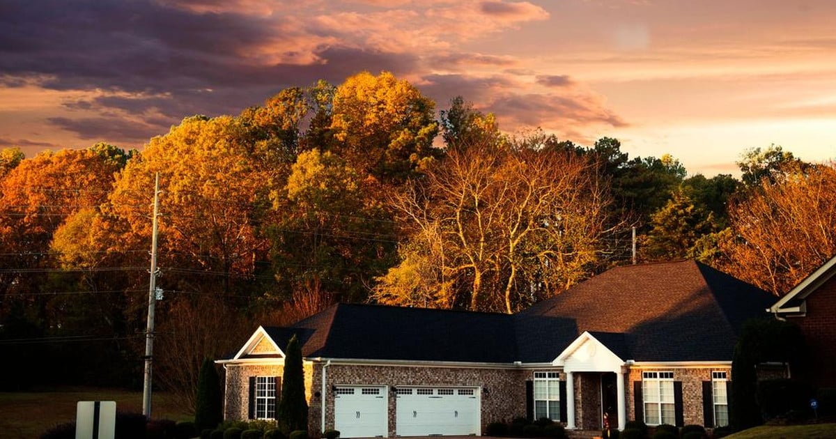 A brick house with a two-car garage on a wooded suburban street at sunset, backed by mature hardwood trees.
