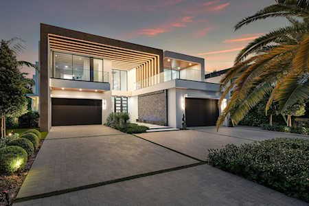 Modern two-story home with illuminated geometric facade, two garage doors, and palm landscaping at dusk.