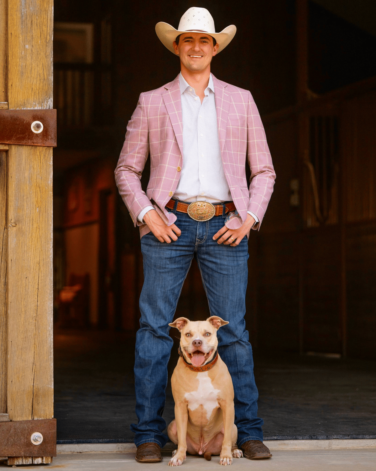 Cody Inman stands in a wooden doorway with a tan dog, wearing a white cowboy hat and pink plaid jacket.