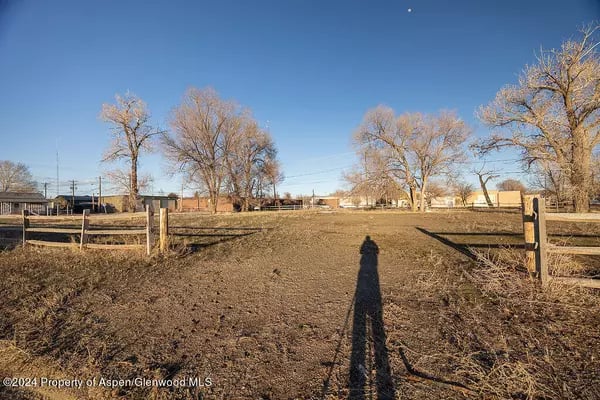 Bare dirt lot bordered by split-rail fencing, with leafless trees and buildings in the distance.