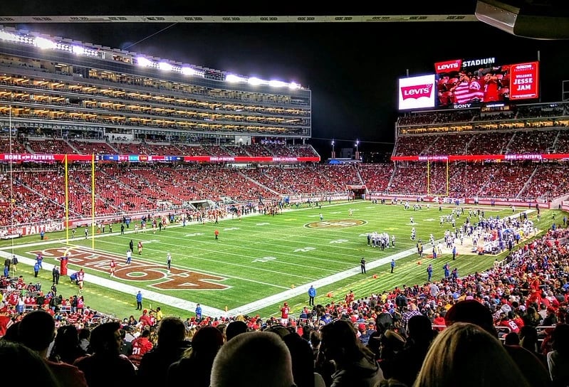 Nighttime view inside Levi&rsquo;s Stadium in Santa Clara with a packed crowd, bright field lights, and players warming up on the field.