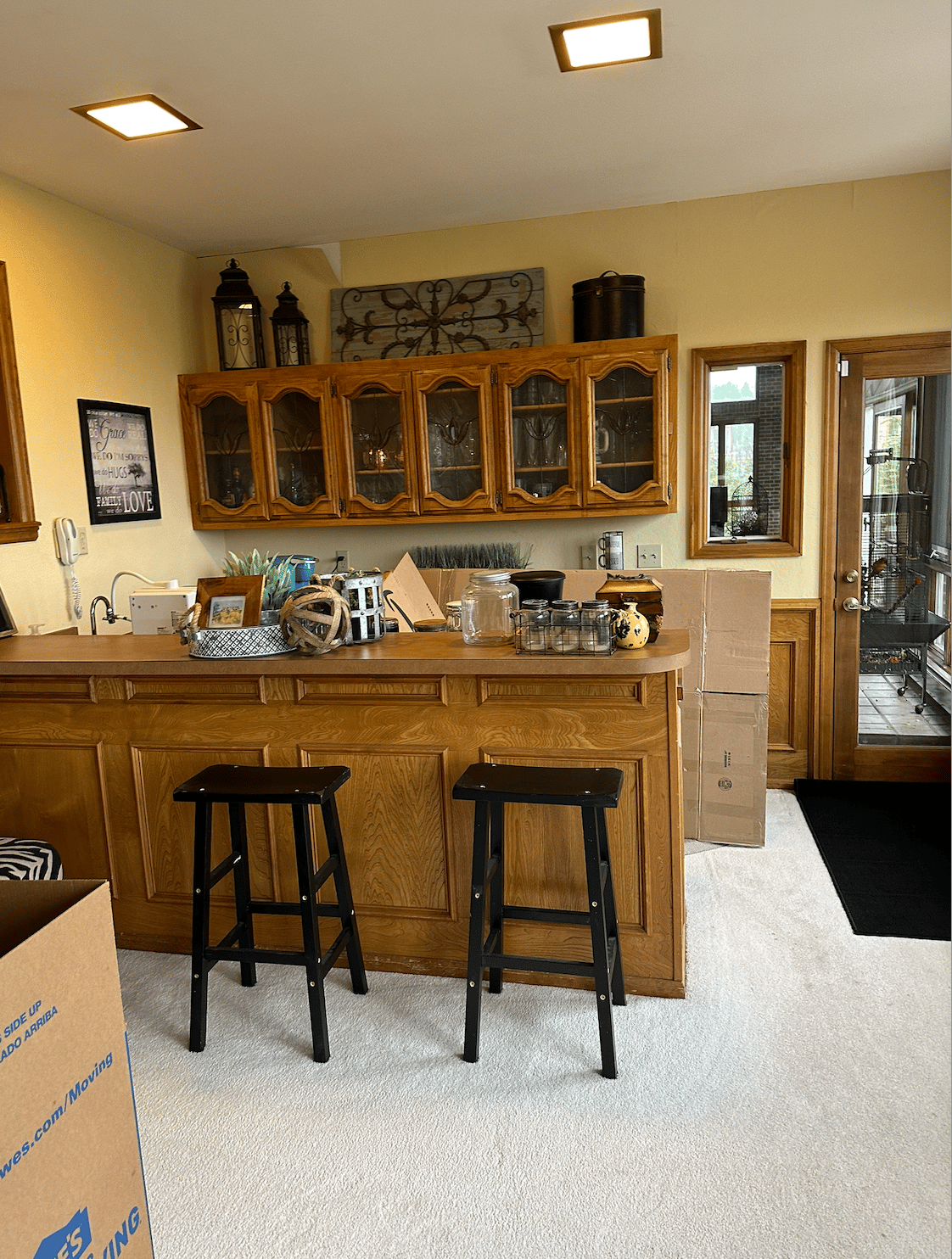 Interior with a wooden bar, glass-front cabinets, two black stools, and moving boxes.