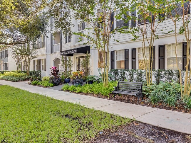 Residential building with balconies and dark shutters, fronted by landscaped gardens and a sidewalk bench.