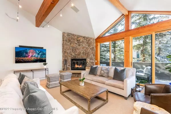 Living room with a stone fireplace, exposed timber beams, and expansive windows overlooking trees.
