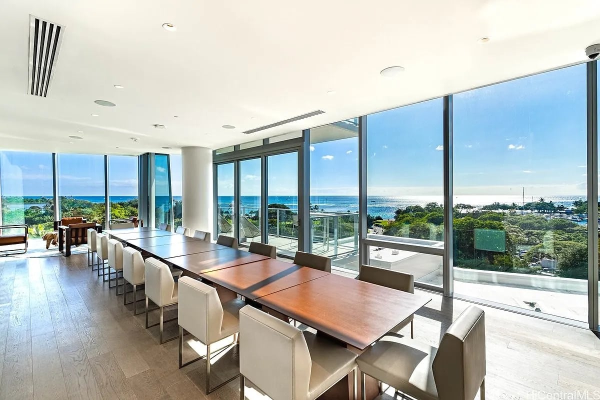 Dining area with a long wood table and cream chairs, overlooking the ocean through floor-to-ceiling windows.