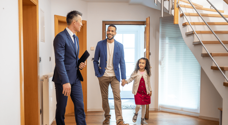 Man in a suit shows a house to a smiling man and a young girl holding hands. They walk through a bright hallway, conveying a sense of excitement.
