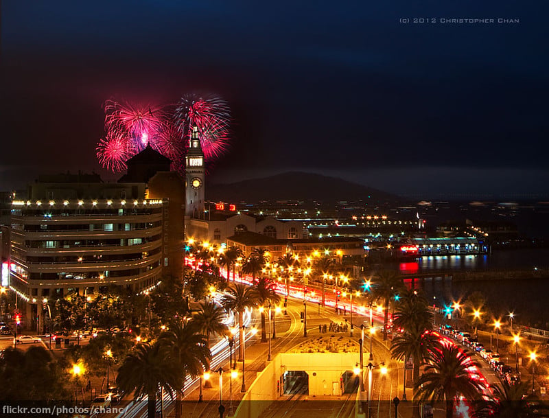 Fourth of July fireworks bursting over the San Francisco Ferry Building at night, with the illuminated Embarcadero waterfront, palm trees, and San Francisco Bay visible below.