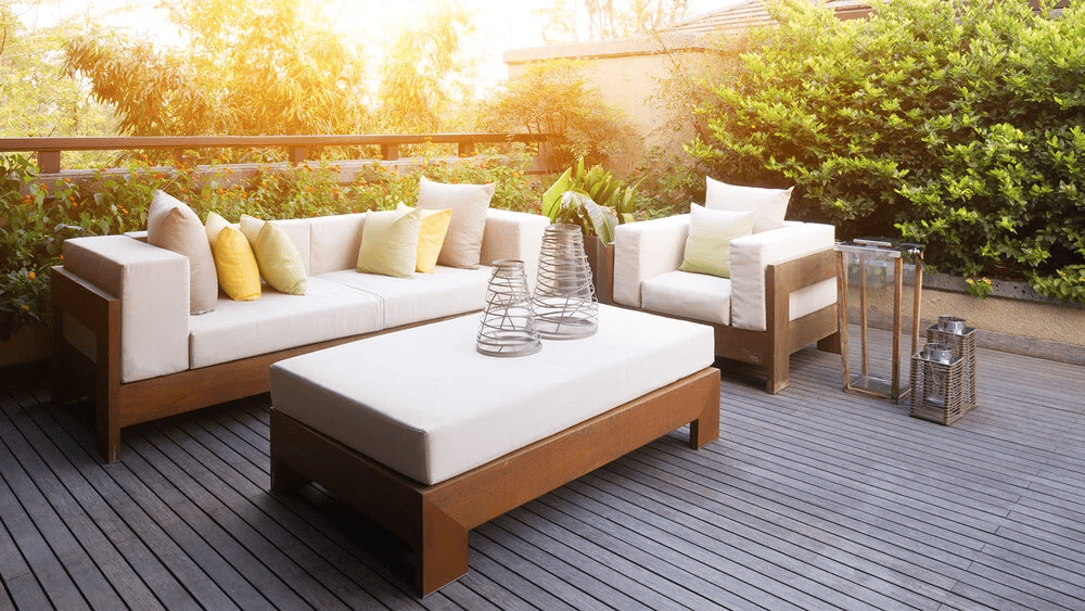 Outdoor deck lounge with white-cushioned wooden seating and greenery in warm sunlight.