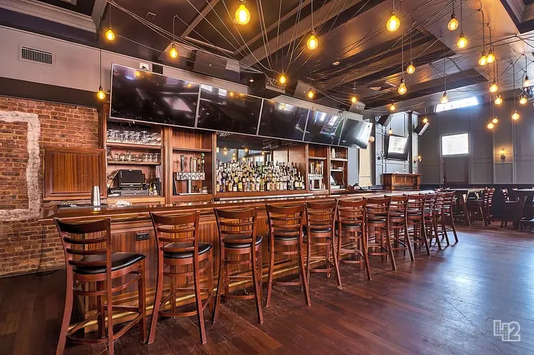 Bar interior with a long wooden counter, rows of stools, and warm hanging lights.