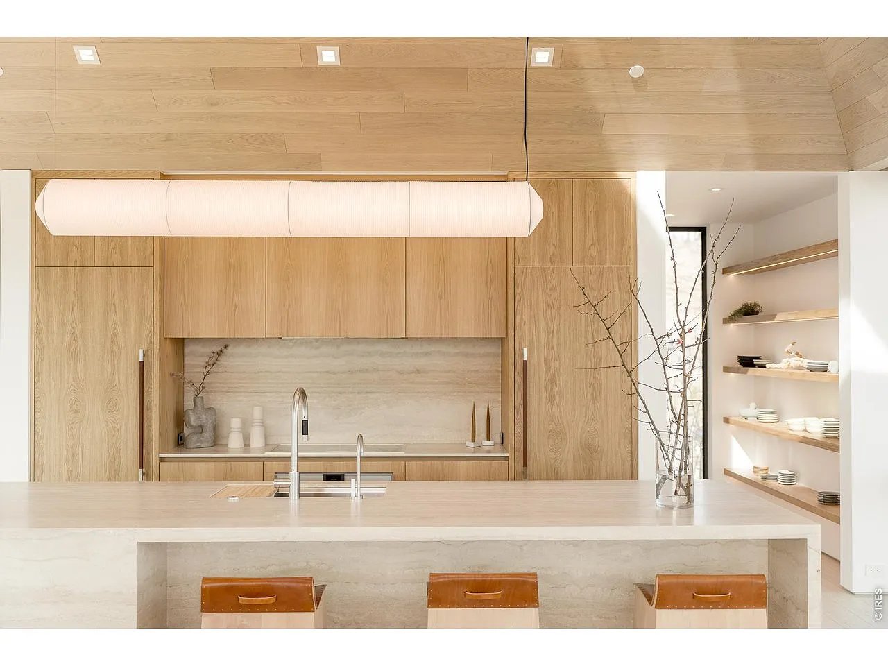 Modern kitchen with light oak cabinetry, a pale stone island, open shelves, and three leather stools.