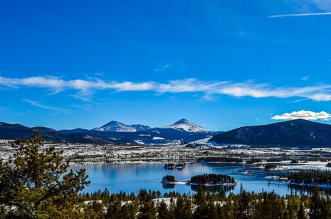Mountain reservoir bordered by snowy peaks and evergreen trees beneath a bright blue sky.