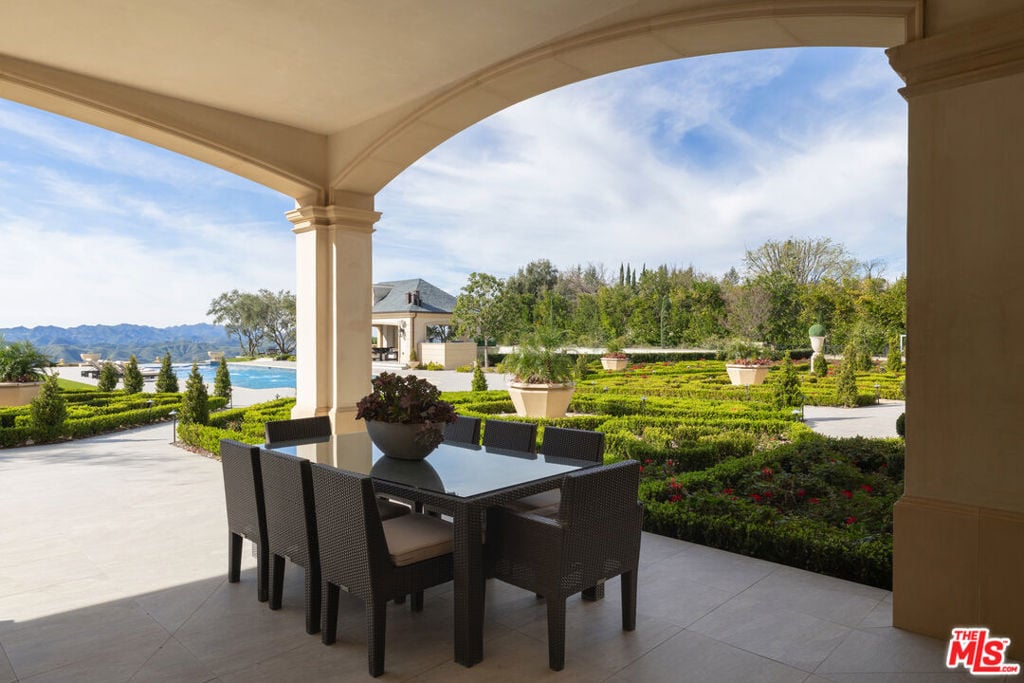Covered patio with dark dining furniture overlooking formal gardens, a swimming pool, and distant mountains.